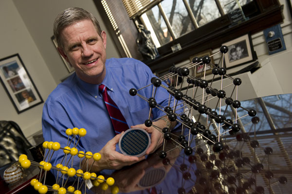 Kevin Hemker, seated between models representing how atoms are packed within an individual grain in a material, holds a silicon wafer onto which nanocrystalline aluminum thin film specimens have been deposited. Photo: Will Kirk/Homewoodphoto.jhu.edu